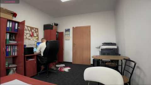 Media: Video of a small, cluttered office with a man in a white shirt working at a desk. Red bookshelves, a white chair, and a messy desk are visible.