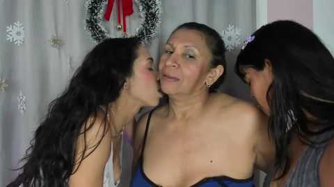 Media: Video of three women in a festive room. Two young women, one in a strapless top, are kissing an older woman with short dark hair, wearing a blue top, in front of a decorated window.