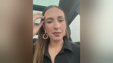 Media: Video of a young woman with fair skin and long, straight brown hair, wearing a black blouse and large, circular earrings, seated in a car. She has a neutral expression and is touching her hair.