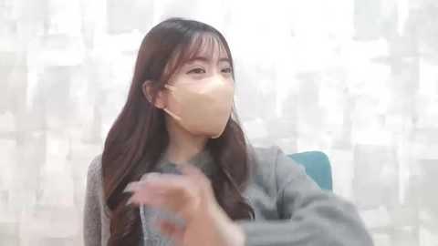 Media: Video of an Asian woman with long black hair, wearing a beige face mask, grey sweater, and a blue chair, against a soft-focus, white abstract background.