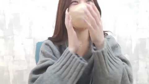 Media: Video of a young East Asian woman with long, straight brown hair, wearing a grey cardigan, covering her mouth with both hands, sitting indoors in a softly lit room with a blurred background.