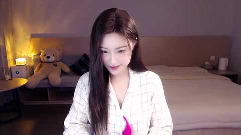 Media: Video of an East Asian woman with long, straight black hair, wearing a white plaid robe, sitting in a dimly lit bedroom with a teddy bear, a lit lamp, and a neatly made bed.