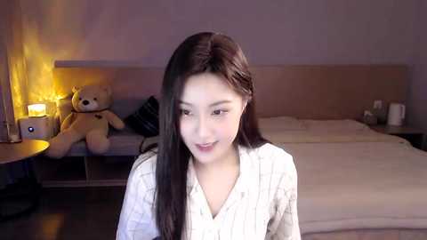 Media: Video of an Asian woman with long, straight black hair, wearing a white, textured robe, seated in a dimly lit bedroom with a beige bed and stuffed toys.