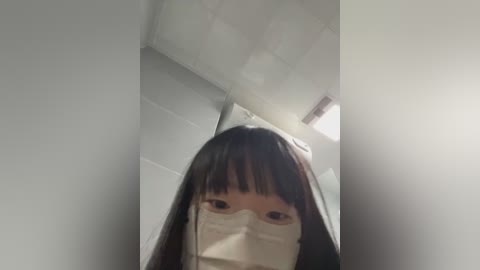 Media: A video shows a young woman with long black hair, wearing a white face mask, standing in a dimly lit room with white walls and a ceiling light.