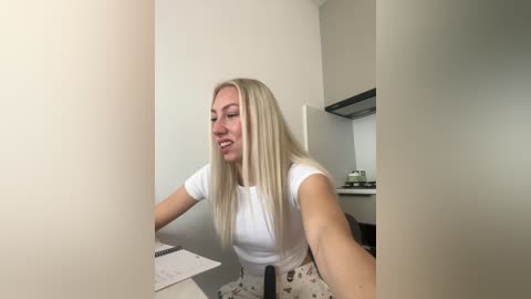 Media: Video of a smiling, blonde, Caucasian woman with straight hair, wearing a white T-shirt and floral-patterned pants, seated in a modern, minimalist kitchen with light-colored walls and stainless steel appliances.