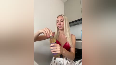 Media: Video of a blonde woman in a red tank top, pouring liquid from a purple bottle into a glass. Minimalist, modern interior with white walls and a flat-screen TV in the background.