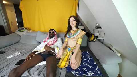 Media: Video of a young white woman in Egyptian-inspired attire, sitting on a blue-patterned blanket next to a Black man in a white robe and pink hijab, in a modern living room with a yellow curtain.