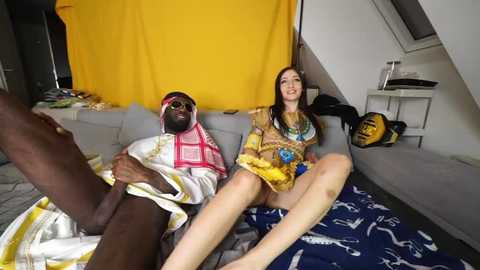 Media: Video of a dark-skinned man in traditional attire and a white woman in a colorful dress, sitting on a bed with a blue and white patterned blanket, in a modern, loft-style room with a yellow curtain.