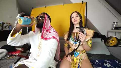 Media: Video: A Black man in a white robe with a red and white headscarf, sunglasses, and a blue can, sits next to a smiling White woman in a yellow and blue dress, both drinking from cans in a messy room with yellow backdrop.