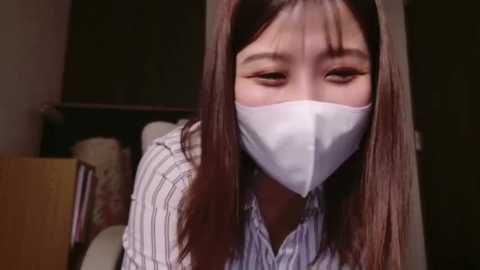 Media: Video of an Asian woman with long brown hair, wearing a white face mask and a striped shirt, indoors, dim lighting.