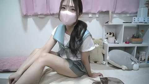 Media: Video of an East Asian woman in a Sailor Moon school uniform, wearing a pink mask, seated on a bed. Background includes a pink curtain, stuffed toys, and a white bookshelf.