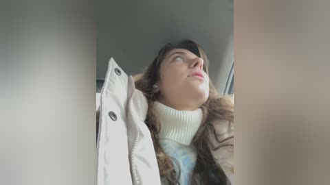 Media: Video of a Caucasian woman with long brown hair, wearing a beige jacket and a white turtleneck, looking upward in a car, with blurred window and seat visible.