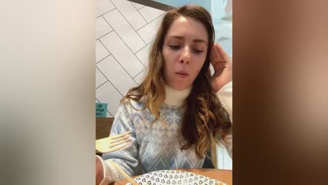 Media: A video of a young woman with long brown hair, wearing a patterned sweater, sitting at a table, looking tired. The background shows a tiled wall and a wooden surface.