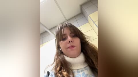 Media: Video of a young woman with long, wavy brown hair, wearing a cream turtleneck sweater, standing in a modern, tiled hallway with fluorescent lighting.