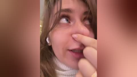 Media: A close-up video of a young woman with light skin, brown hair, and blue eyes, wearing a white turtleneck sweater. She has a shocked expression, with her hand covering her mouth.