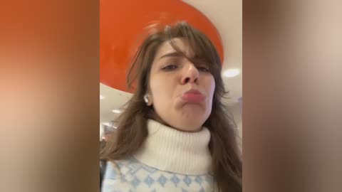 Media: Video of a young woman with fair skin, brown hair, and a white turtleneck sweater, making a pouty face indoors against a bright orange wall.