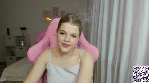 Media: A video of a young woman with light skin and straight brown hair in a pink gaming chair, wearing a white spaghetti-strap tank top, in a softly lit bedroom with white curtains.