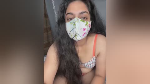 Media: Video of a young woman with long, dark hair, wearing a floral-patterned bra and a face mask, sitting in a dimly lit room with a chain hanging in the background.