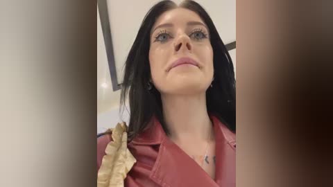 Media: A close-up video of a woman with long black hair, fair skin, and blue eyes, wearing a red jacket with gold accents, standing indoors.