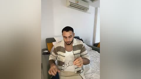 Media: A video of a bearded man in a striped cardigan, sitting on a bed in a minimalist, well-lit room with white walls, an air conditioner, and a gray sofa in the background.