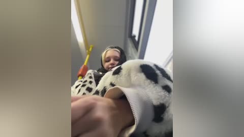 Media: A video captures a young woman with long blonde hair, wearing a black-and-white cow-patterned sweater, leaning into a yellow and red handrail in a dimly lit subway car.