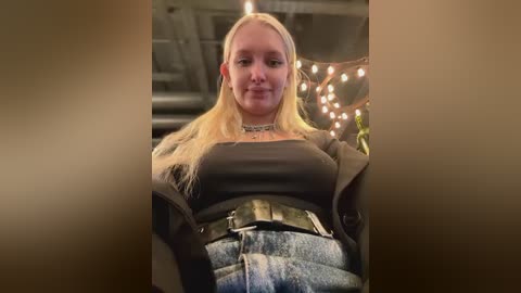 Media: A video of a blonde woman wearing a black crop top and denim jeans, smiling with a choker necklace, in a dimly-lit room with warm string lights and a staircase background.