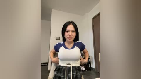 Media: Video of a young woman with long black hair, wearing a white and blue Adidas shirt and a plaid skirt, standing in a minimalistic, modern room with white walls and a wooden floor.