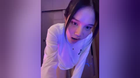 Media: A video of a young Asian woman with straight black hair and light skin, wearing a white top, bending forward with her hand on her cheek, in a dimly lit room with purple lighting.