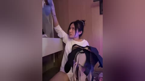 Media: Video of an Asian woman with dark hair in a bun, wearing a white sweater, sitting on the floor, reaching for a box in a dimly lit room with purple lighting.