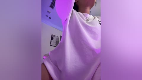 Media: Video of a woman in a white shirt, standing in a dimly lit room with purple lighting. She has light skin and is lifting her shirt to expose her stomach. The background features a white wall with framed black-and-white artwork.