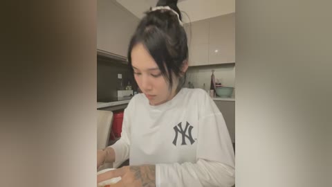 Media: Video of an Asian woman with dark hair in a bun, wearing a white New York Yankees sweatshirt, cooking in a modern kitchen with beige cabinets and a red countertop.