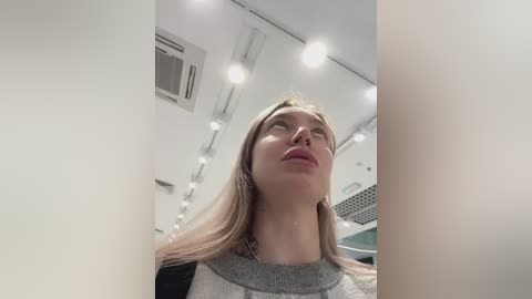 Media: Video of a young Caucasian woman with long, straight blonde hair, wearing a grey sweater, looking upward, inside a modern, white interior with hanging lights and an air conditioning unit in the background.