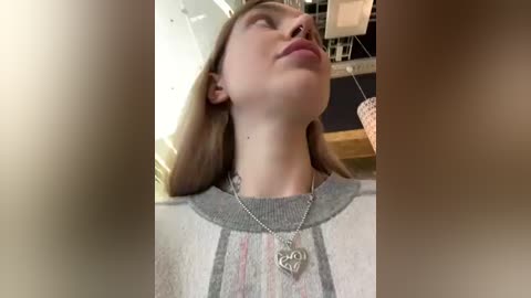 Media: Video of a young woman with light skin and straight, shoulder-length brown hair, wearing a gray sweater with a heart pendant necklace, indoors with blurred furniture and lighting fixtures.