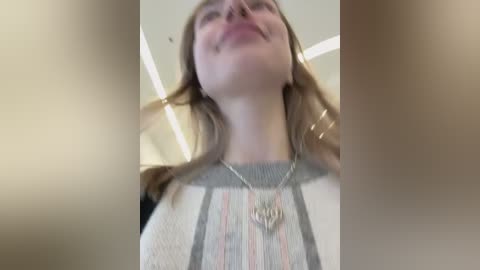 Video of a smiling Caucasian woman with light skin, wearing a striped sweater, silver necklace, and brown hair, captured from a low angle, with blurred background.