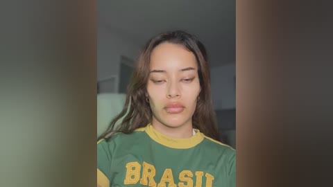 Video of a young woman with light brown skin, long dark hair, and a neutral expression, wearing a green and yellow Brazilian soccer jersey. Background is blurred and neutral-toned.