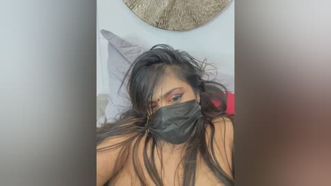 Media: Video of a topless, dark-skinned woman with long, dark hair, wearing a black face mask, and a red top, lying on a bed with a brown circular wall decoration in the background.