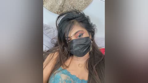 Media: Video of a young woman with long black hair, wearing a black face mask, revealing her orange eyeshadow and teal lace bralette. She is indoors, with a beige wall and a circular patterned wall hanging.