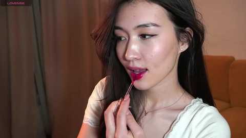 Media: Video of an Asian woman with long black hair, wearing a white top, sensually licking a red lollipop, in a softly lit room with beige curtains and a brown couch.