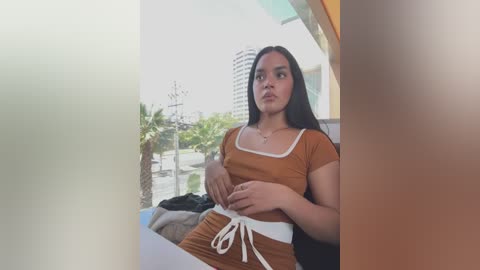 Media: Video of a young Latina woman with long dark hair, medium skin tone, and medium build, wearing a brown crop top and shorts, sitting on a balcony with a cityscape background.