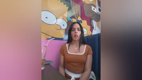 Media: Video of a young woman with long black hair, wearing a brown crop top and white skirt, sitting at a table. Background features colorful cartoon characters and a pink drink.