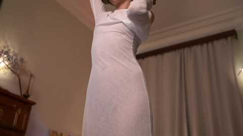 Media: A video of a woman in a sheer white dress, standing indoors with beige walls, a wooden cabinet, and a white curtain in the background.