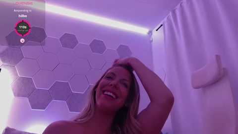 Media: Video of a smiling, topless blonde woman with fair skin, wearing a white towel, in a modern, purple-lit bathroom with hexagonal tile and a white shower curtain.