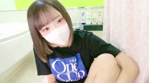 Media: Video of an Asian woman with straight brown hair, wearing a blue T-shirt with white text, a white mask, and sitting in a white-tiled bathroom.