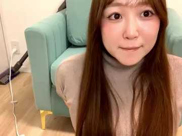 Media: Video of a young Asian woman with long, straight brown hair and light skin, wearing a beige turtleneck, sitting on a light teal couch in a modern, minimalist room with wooden floors and white walls.