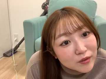 Media: Video of a young East Asian woman with long, straight brown hair and fair skin, wearing a light grey sweater, sitting on a teal armchair in a modern, minimalist room with light wooden flooring.