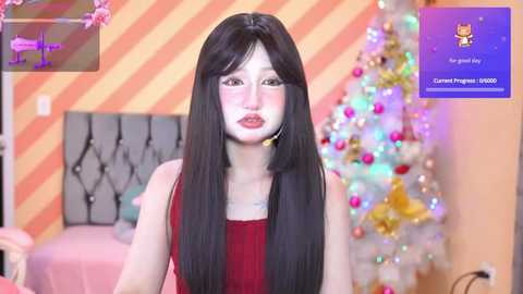 Media: A video of a young East Asian woman with long black hair, fair skin, and red lipstick, dressed in a red dress, standing in front of a Christmas tree with colorful ornaments in a warmly lit room.