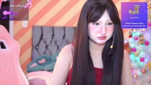 Media: Video of a young Asian woman with long black hair and fair skin, wearing a red dress, sitting in a pink gaming chair in a brightly colored, cartoon-themed room with a Christmas tree.