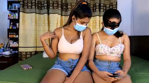 Media: Video of two young women with light brown skin, wearing face masks and matching light blue crop tops, sitting on a green bed in a bedroom with patterned curtains and a bookshelf.