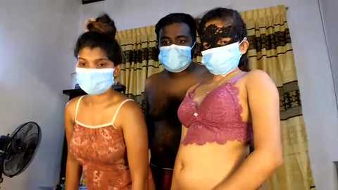 Media: Video of three individuals in a room, wearing light blue masks, two in lace lingerie, one in a patterned dress, standing in front of a yellow curtain.