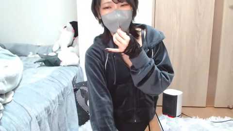 Media: Video of an Asian woman in a gray face mask, dark hair, and black hoodie, standing in a cluttered bedroom with a gray bed, wooden closet, and white trash can.